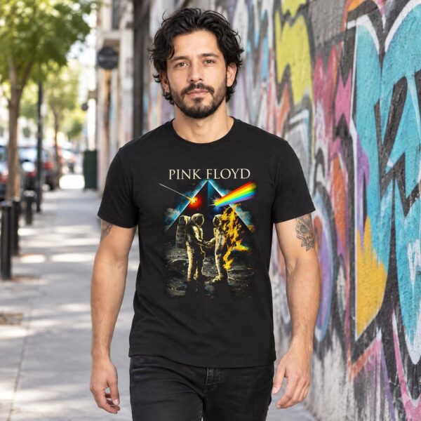 Hombre con playera negra Pink Floyd de astronautas y prisma caminando junto a un muro con grafiti
